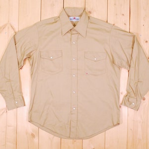 May include: A light brown long-sleeved button-down shirt with two chest pockets and a single button cuff. The shirt has a white button placket and a small red logo on the left chest pocket.