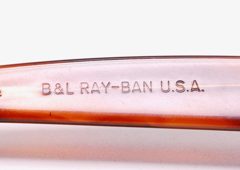 Puede incluir: Primer plano del brazo de pl&aacute;stico marr&oacute;n de unas gafas de sol Ray-Ban. El texto "B&L RAY-BAN U.S.A." est&aacute; grabado en relieve en el brazo.