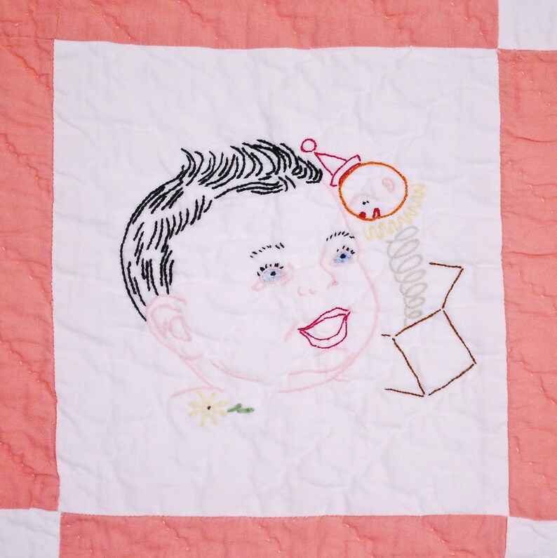 May include: Embroidered quilt square featuring a smiling baby with black hair and a toy jack-in-the-box with a clown face.