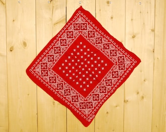 Pode incluir: Bandana vermelha com um padrão paisley branco e pontos pretos. A bandana está dobrada em forma de triângulo e pendurada em uma parede de madeira.