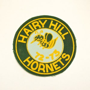 Pode incluir: Etiqueta bordada verde e amarela com o texto "Hairy Hill Hornets" e os anos "72-73" em um círculo. Uma abelha estilizada está no centro do círculo.