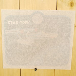 Puede incluir: Una impresión rectangular de Star Trek, parcialmente cubierta por una hoja de papel translúcido. Las palabras "STAR TREK" son visibles en letras blancas en la parte superior. La imagen de abajo parece representar la nave espacial Enterprise.