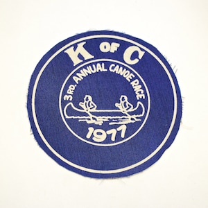 Może przedstawiać: Niebieska okrągła naszywka z białym tekstem i białą sylwetką dwóch osób w kajaku. Tekst brzmi "K of C 3rd. Annual Canoe Race 1971".