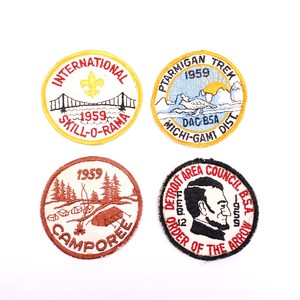 Könnte beinhalten: Vier runde gestickte Aufnäher mit verschiedenen Designs. Der obere linke Aufnäher ist gelb mit roter und schwarzer Schrift, die "INTERNATIONAL 1959 SKILL-O-RAMA" lautet. Der obere rechte Aufnäher ist gelb mit roter und schwarzer Schrift, die "PTARMIGAN TREK 1959 DAC-B5A MICHI-GAMI DIST" lautet. Der untere linke Aufnäher ist braun mit weißer Schrift, die "1959 CAMPOREE" lautet. Der untere rechte Aufnäher ist schwarz mit roter und weißer Schrift, die "DETROIT AREA COUNCIL BSA 1959 ORDER OF THE ARROW" lautet.