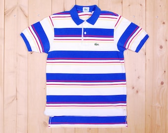 Könnte beinhalten: Ein blau-weiß gestreiftes Poloshirt mit roten und gelben Streifen. Das Shirt hat ein kleines grünes Krokodil-Logo auf der linken Brust.