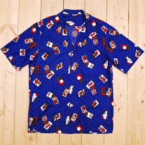 Puede incluir: Una camisa azul de manga corta con botones y un diseño estampado en blanco y rojo. El estampado presenta varias formas geométricas y símbolos.