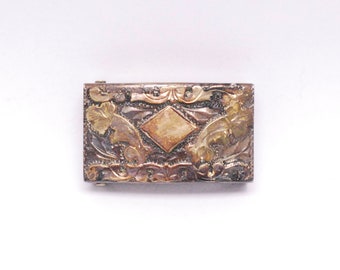 May include: A silver belt buckle with a diamond-shaped design in the center and floral patterns on the sides. The buckle is made of metal and has a polished finish.