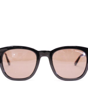 Może przedstawiać: Czarne okulary przeciwsłoneczne z brązowymi szkłami i złotymi akcentami. Okulary mają napis "TOM FORD" nadrukowany na prawym szkle.