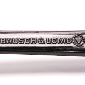 以下が含まれることがあります： 黒い金属フレームのクローズアップ。フレームには「BAUSCH & LOMB」の文字と三角形のロゴが刻まれています。
