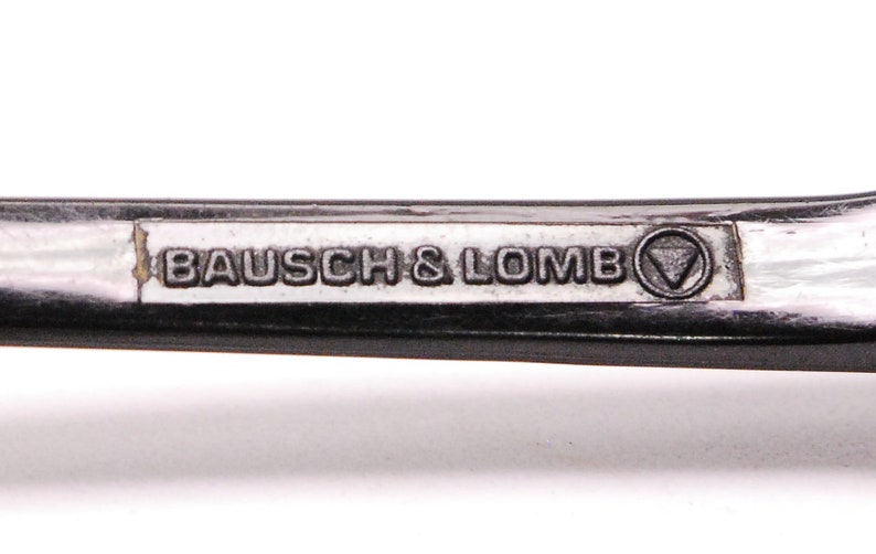以下が含まれることがあります： 黒い金属フレームのクローズアップ。フレームには「BAUSCH & LOMB」の文字と三角形のロゴが刻まれています。
