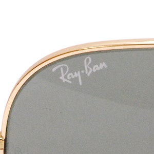 Pode incluir: Close-up de uma lente cinza com uma arma&ccedil;&atilde;o dourada. O logotipo Ray-Ban est&aacute; gravado na lente.