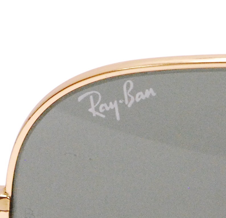 Pode incluir: Close-up de uma lente cinza com uma arma&ccedil;&atilde;o dourada. O logotipo Ray-Ban est&aacute; gravado na lente.