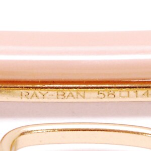 Pode incluir: Close-up da arma&ccedil;&atilde;o de metal dourado de um par de &oacute;culos de sol Ray-Ban. A arma&ccedil;&atilde;o tem o texto "RAY-BAN 58 114" gravado.