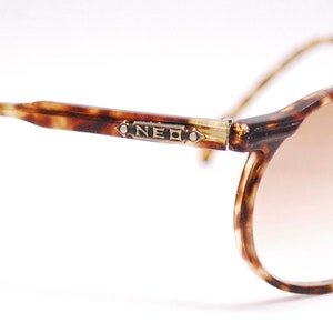 May include: Tortoiseshell frame sunglasses with brown gradient lenses. The sunglasses have a metal accent on the bridge of the frame with the word "NEO" engraved on it.