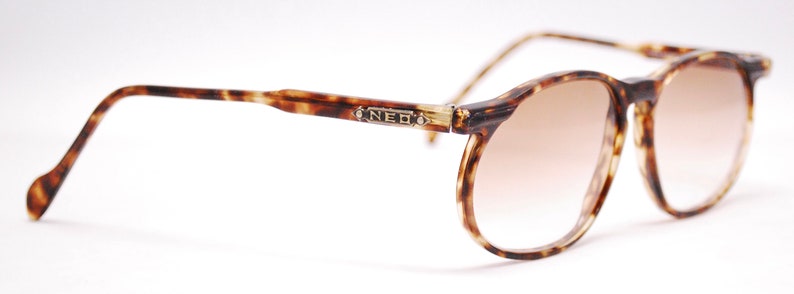 May include: Tortoiseshell frame sunglasses with brown gradient lenses. The sunglasses have a metal accent on the bridge of the frame with the word "NEO" engraved on it.