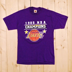 Puede incluir: Camiseta morada con el logotipo de los Los Angeles Lakers y el texto "1985 NBA CHAMPIONS" impreso en la parte delantera.
