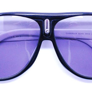 Puede incluir: Gafas de sol aviador negras con lentes morados. Las gafas tienen un puente de metal plateado y el texto "CARRERA 5200/S 99/IR 135 Made in Italy" est&aacute; impreso en la patilla derecha.