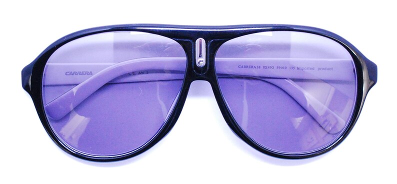 Puede incluir: Gafas de sol aviador negras con lentes morados. Las gafas tienen un puente de metal plateado y el texto "CARRERA 5200/S 99/IR 135 Made in Italy" est&aacute; impreso en la patilla derecha.