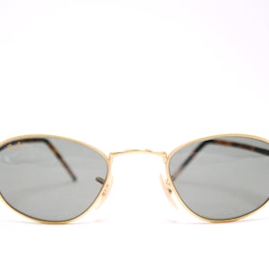 May include: Gold-rimmed sunglasses with dark gray lenses. The sunglasses have a thin, oval frame.