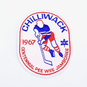 Puede incluir: Un parche ovalado blanco con bordados rojos y azules. Presenta a un jugador de hockey patinando con un palo y un disco. El texto dice "Chilliwack 1967 Centennial Pee Wee Jamboree".