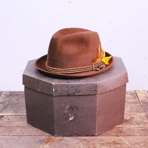 Op de afbeelding: Een bruine fedora hoed met een bruin touwband en gele veren bovenop. De hoed ligt op een bruine doos.