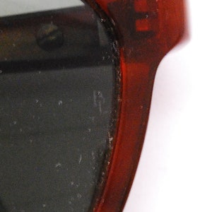 May include: Close-up of the red plastic frame of a pair of sunglasses. The lenses are dark and the frame has a slight curve.