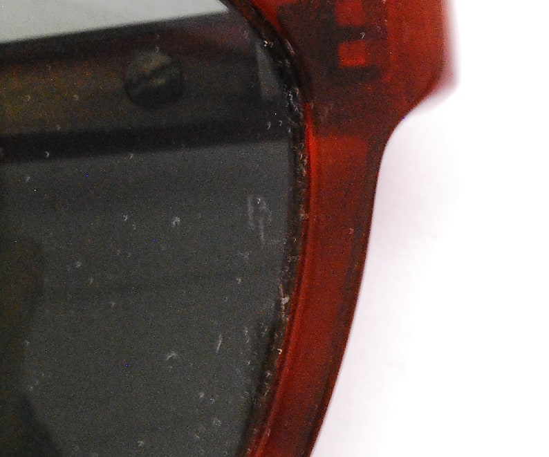 May include: Close-up of the red plastic frame of a pair of sunglasses. The lenses are dark and the frame has a slight curve.