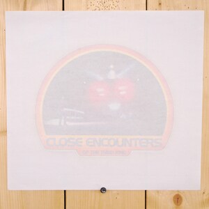 May include: A print of a movie poster for "Close Encounters of the Third Kind" is displayed on a wooden surface. The poster features a colorful design with the film's title and imagery of a spaceship and red lights.