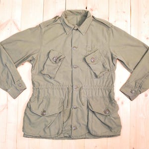 May include: An olive green military-style jacket with a button-down front, two chest pockets, and two lower pockets. The jacket has long sleeves with buttoned cuffs and a drawstring waist. The collar is a classic point collar.