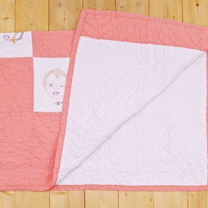 May include: A pink and white quilt with a simple embroidered design of a child's face. The quilt is folded in half, showing both sides.
