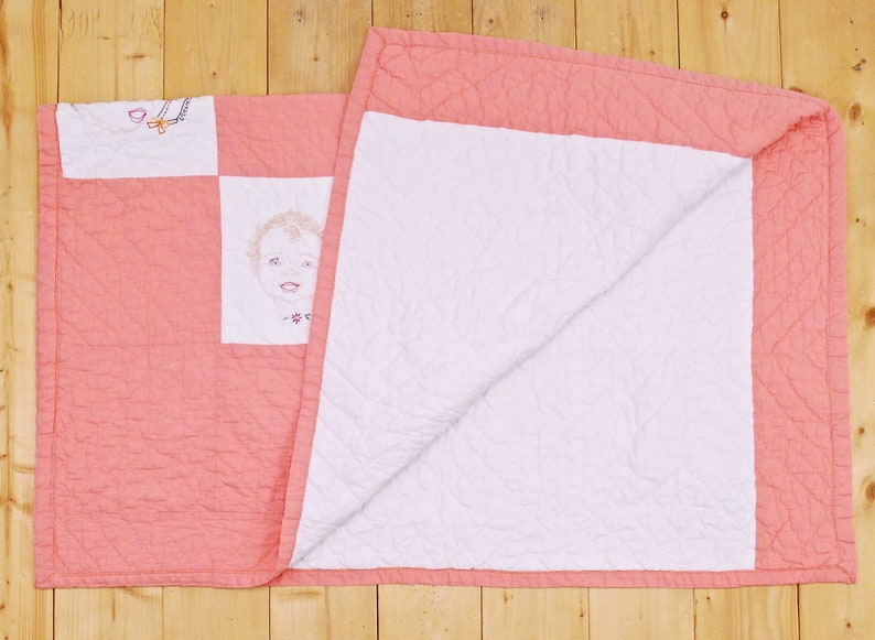 May include: A pink and white quilt with a simple embroidered design of a child's face. The quilt is folded in half, showing both sides.