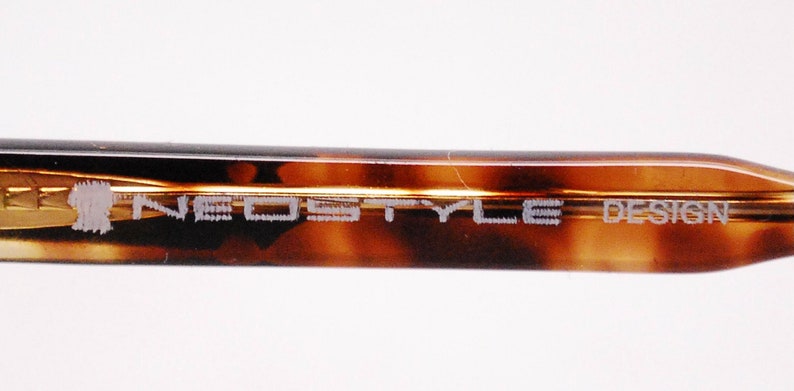 May include: Close-up of the brown tortoiseshell temple arm of a pair of spectacles. The arm is engraved with the text "NEO STYLE DESIGN".