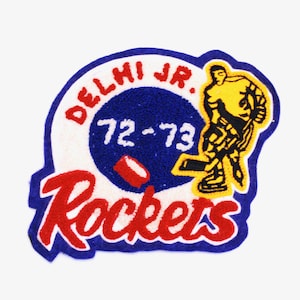 Puede incluir: Un parche de fieltro azul y blanco con el texto "Delhi Jr. 72-73 Rockets" en letras rojas y blancas. Un jugador de hockey de dibujos animados con un jersey amarillo sostiene un palo de hockey y un disco.