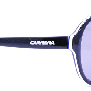 Puede incluir: Gafas de sol aviador negras con un detalle de banda blanca y azul en el marco. Las gafas de sol llevan el logotipo de Carrera en el marco.