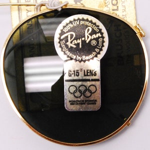 May include: Round sunglasses with dark green lenses and a gold rim. The sunglasses have a sticker that says "100% UV protection Ray-Ban Sunglasses by Bausch & Lomb" and another sticker that says "G-15 Lens" with the Olympic rings and "Worldwide Sponsor 1980 Olympic Games".