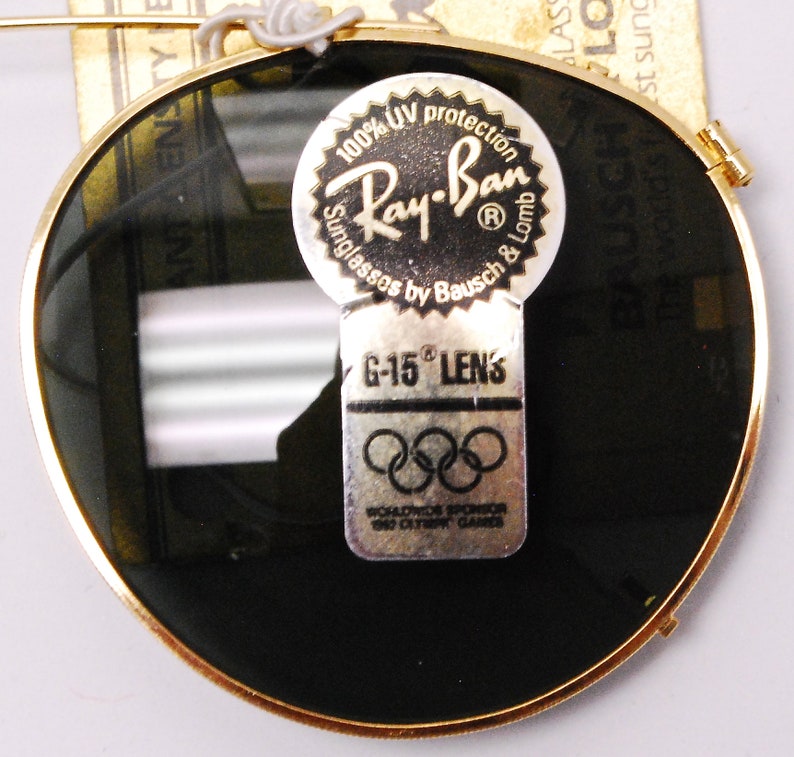 May include: Round sunglasses with dark green lenses and a gold rim. The sunglasses have a sticker that says "100% UV protection Ray-Ban Sunglasses by Bausch & Lomb" and another sticker that says "G-15 Lens" with the Olympic rings and "Worldwide Sponsor 1980 Olympic Games".