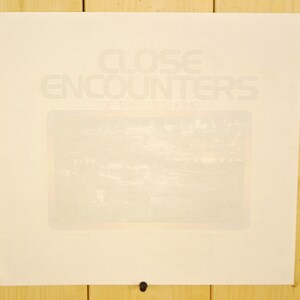 May include: A cream-coloured poster with the words "CLOSE ENCOUNTERS" in a light grey font. Below the title is the text "OF THE THIRD KIND". The poster is mounted on a wooden surface.