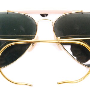 Pode incluir: &Oacute;culos de sol aviador dourados com lentes verdes e um logotipo Ray-Ban na lente direita.