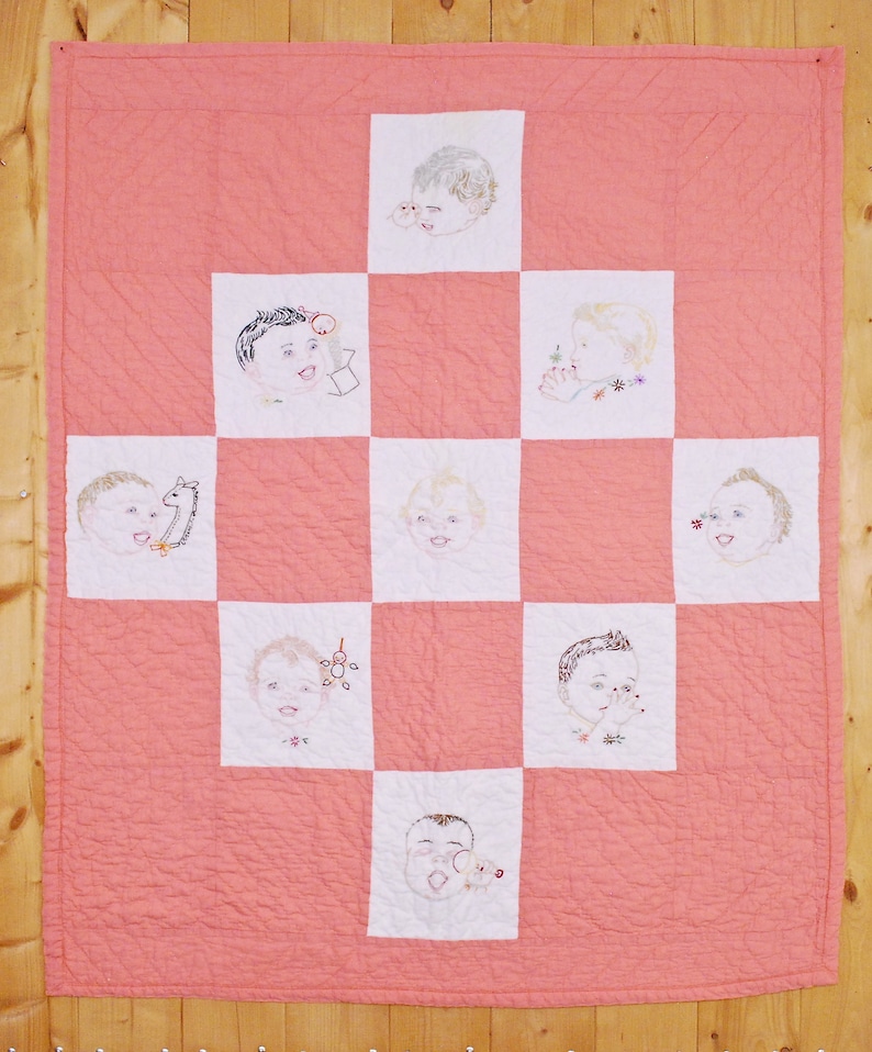 May include: A pink quilt with a white grid pattern. Each square features an embroidered image of a baby's face.