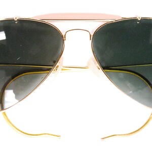 Pode incluir: &Oacute;culos de sol aviador dourados com lentes verdes e um logotipo Ray-Ban na lente esquerda.