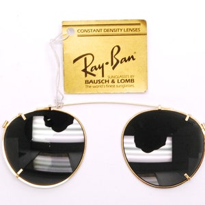 May include: Gold Ray-Ban clip-on sunglasses with black lenses. The sunglasses are attached to a gold metal frame. The tag reads "CONSTANT DENSITY LENSES Ray-Ban SUNGLASSES BY BAUSCH & LOMB The world's finest sunglasses."