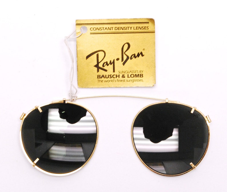 May include: Gold Ray-Ban clip-on sunglasses with black lenses. The sunglasses are attached to a gold metal frame. The tag reads "CONSTANT DENSITY LENSES Ray-Ban SUNGLASSES BY BAUSCH & LOMB The world's finest sunglasses."