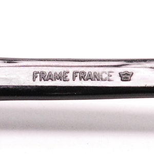 以下が含まれることがあります： 黒いプラスチックフレームのクローズアップ。フレームには「FRAME FRANCE」という文字と王冠のシンボルが刻まれています。