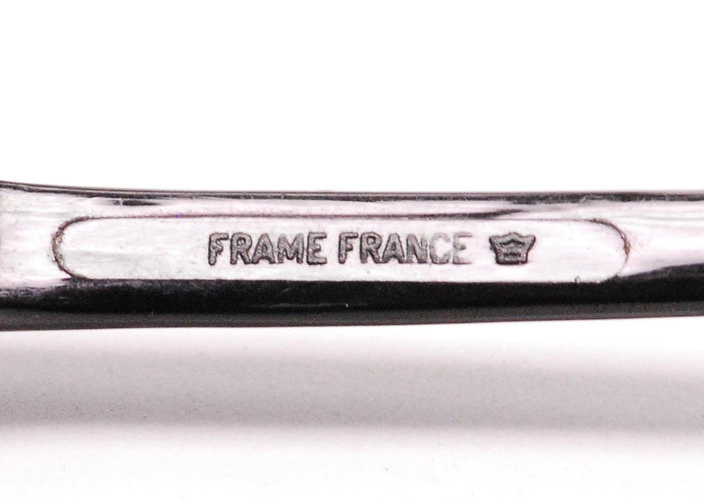 以下が含まれることがあります： 黒いプラスチックフレームのクローズアップ。フレームには「FRAME FRANCE」という文字と王冠のシンボルが刻まれています。