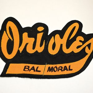 May include: A black and orange felt patch with the word "Orioles" in orange lettering and the words "BAL MORAL" in black lettering.