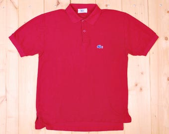May include: A red polo shirt with short sleeves and a collared neckline. The shirt features a two-button placket and a small blue crocodile logo on the left chest. The shirt is made of a soft material.
