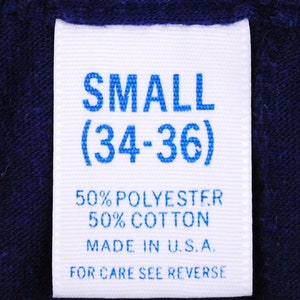 Puede incluir: Etiqueta blanca con texto azul que indica una talla "SMALL (34-36)". La etiqueta tambi&eacute;n dice "50% POLYESTER 50% COTTON MADE IN U.S.A. FOR CARE SEE REVERSE."