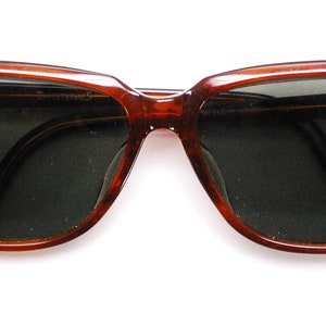 May include: Brown plastic framed sunglasses with dark green lenses. The sunglasses have the Ray-Ban logo on the left frame.
