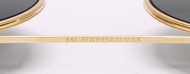 May include: Close-up of the inscription on a pair of gold-tone Ray-Ban sunglasses. The inscription reads "B&L RAY-BAN U.S.A."