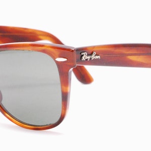 Puede incluir: Gafas de sol Ray-Ban Wayfarer de carey con lentes verdes. Las gafas tienen un logotipo Ray-Ban plateado en la patilla derecha.